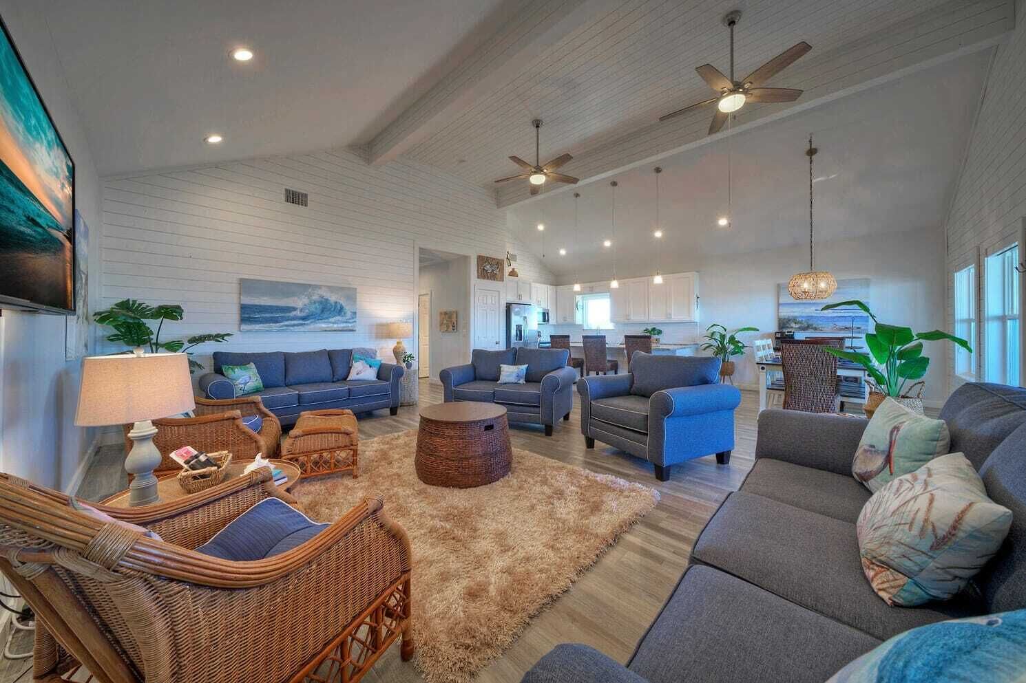 A spacious living room with a large gray sofa, blue armchairs, and a coffee table. There are wicker chairs, a rug, and ceiling fans.