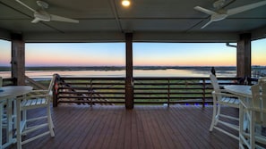 A covered outdoor area with a view of a body of water at sunset, wooden deck, and seating. - Pelican's Porch by Avantstay Private Pool, Views (Port Aransas)