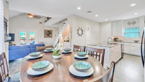 A dining area with a wooden table set for six, surrounded by chairs. A kitchen with white cabinets and a sink is visible in the background. - Makaira Azul by Avantstay Pool & Putting Green! (Port Aransas)