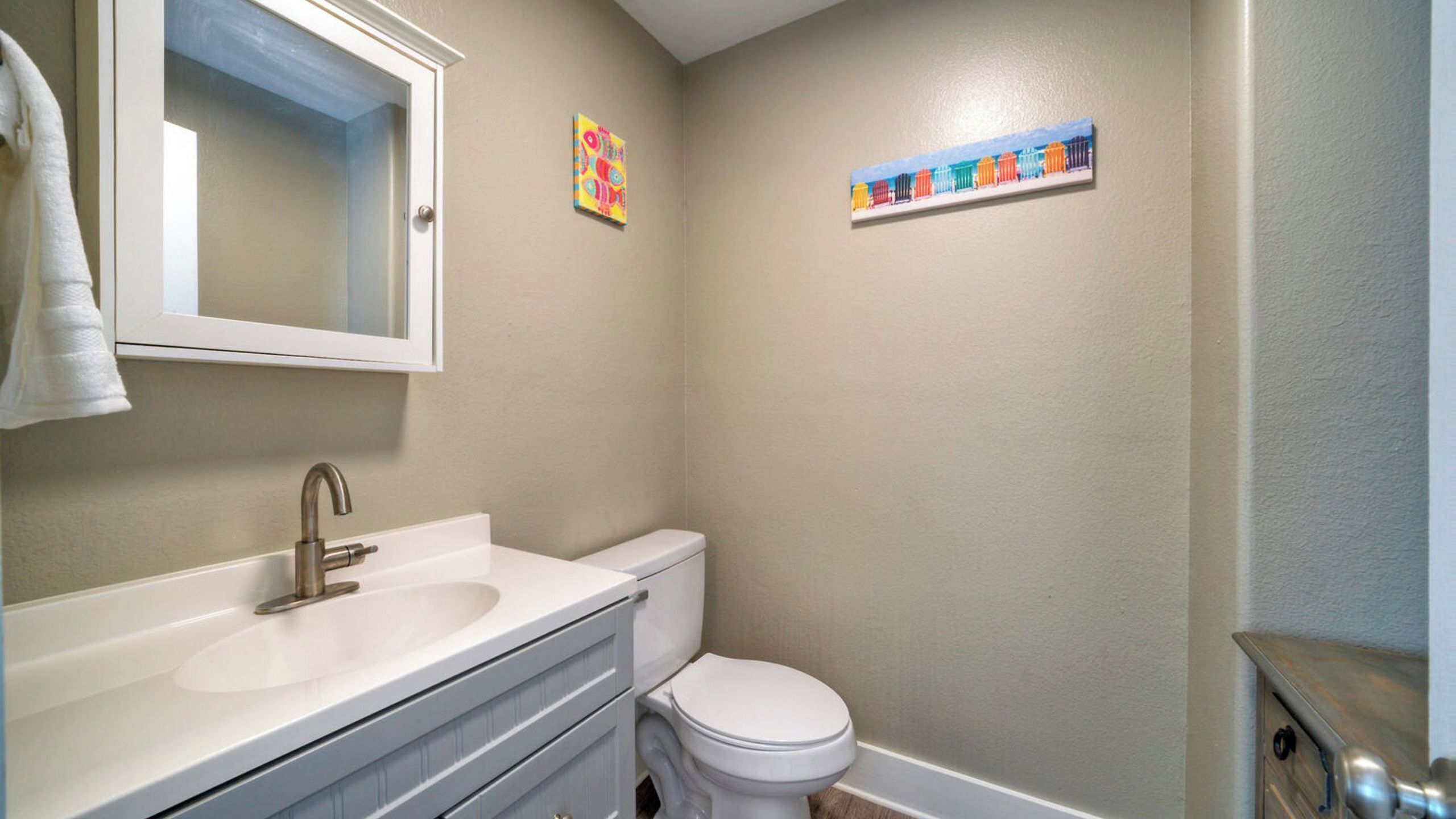 Un bagno con un lavandino bianco, un water e uno specchio sopra il lavandino. In evidenza, due piccoli dipinti alle pareti.