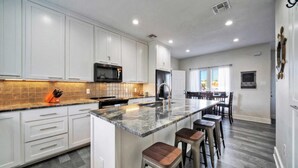 A modern kitchen with white cabinets, a large island with stools, a microwave, and a refrigerator. - Makaira Azul by Avantstay Pool & Putting Green! (Port Aransas)