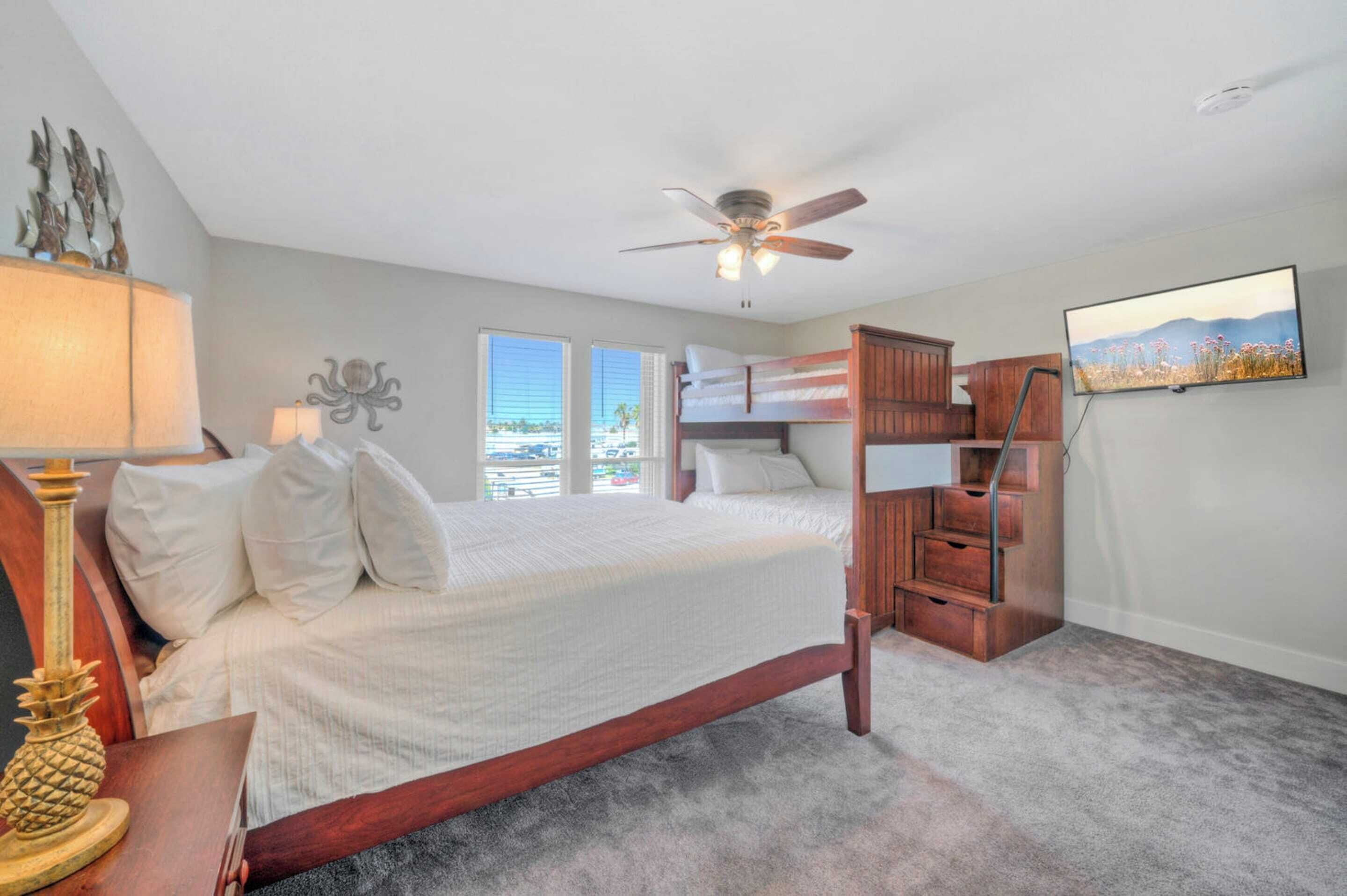 A bedroom with a large bed, a ceiling fan, a wooden staircase, and a TV on the wall.