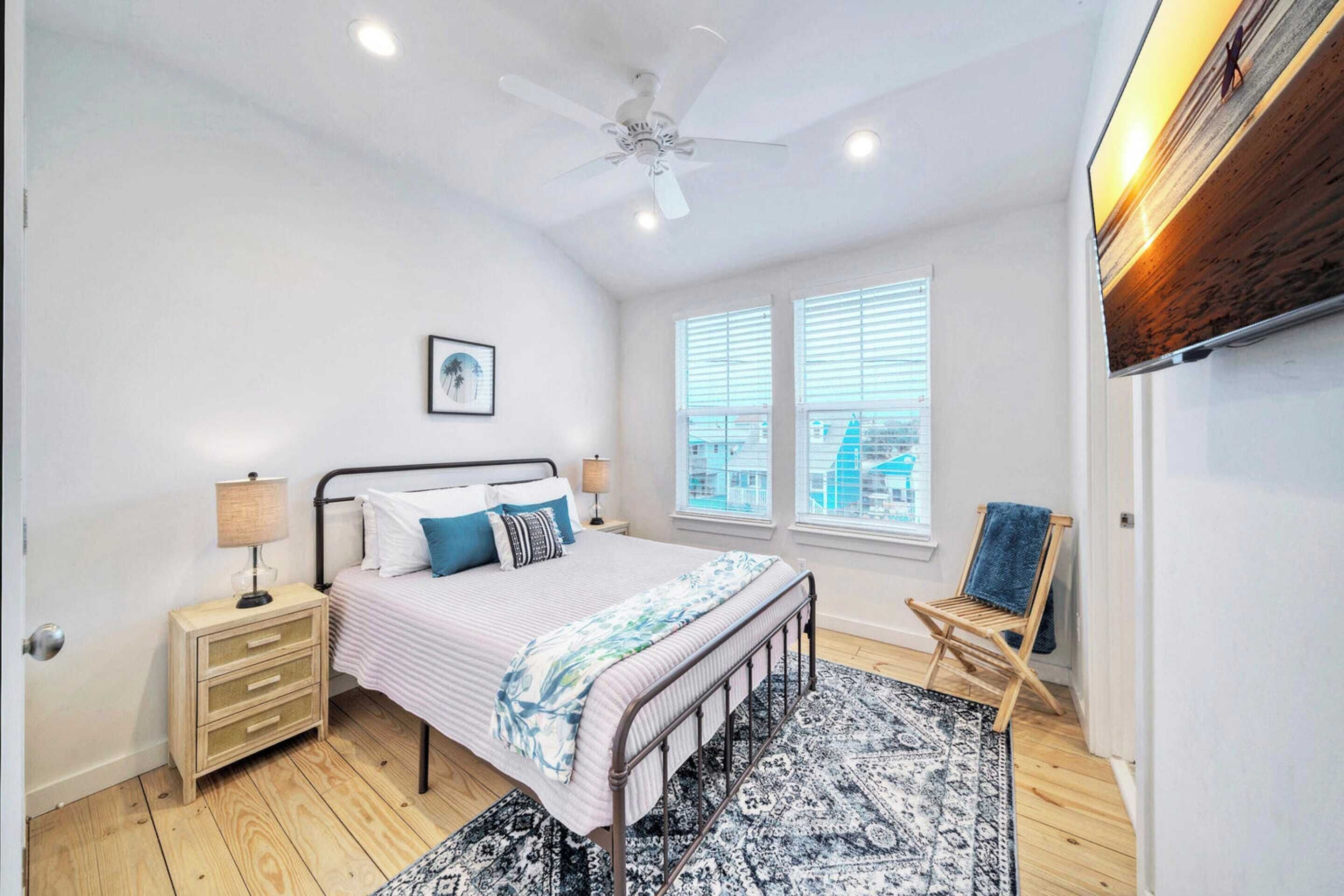 Une chambre à coucher comprenant un lit, des tables de chevet, un ventilateur de plafond, une fenêtre avec des stores, une chaise et un téléviseur fixé au mur.