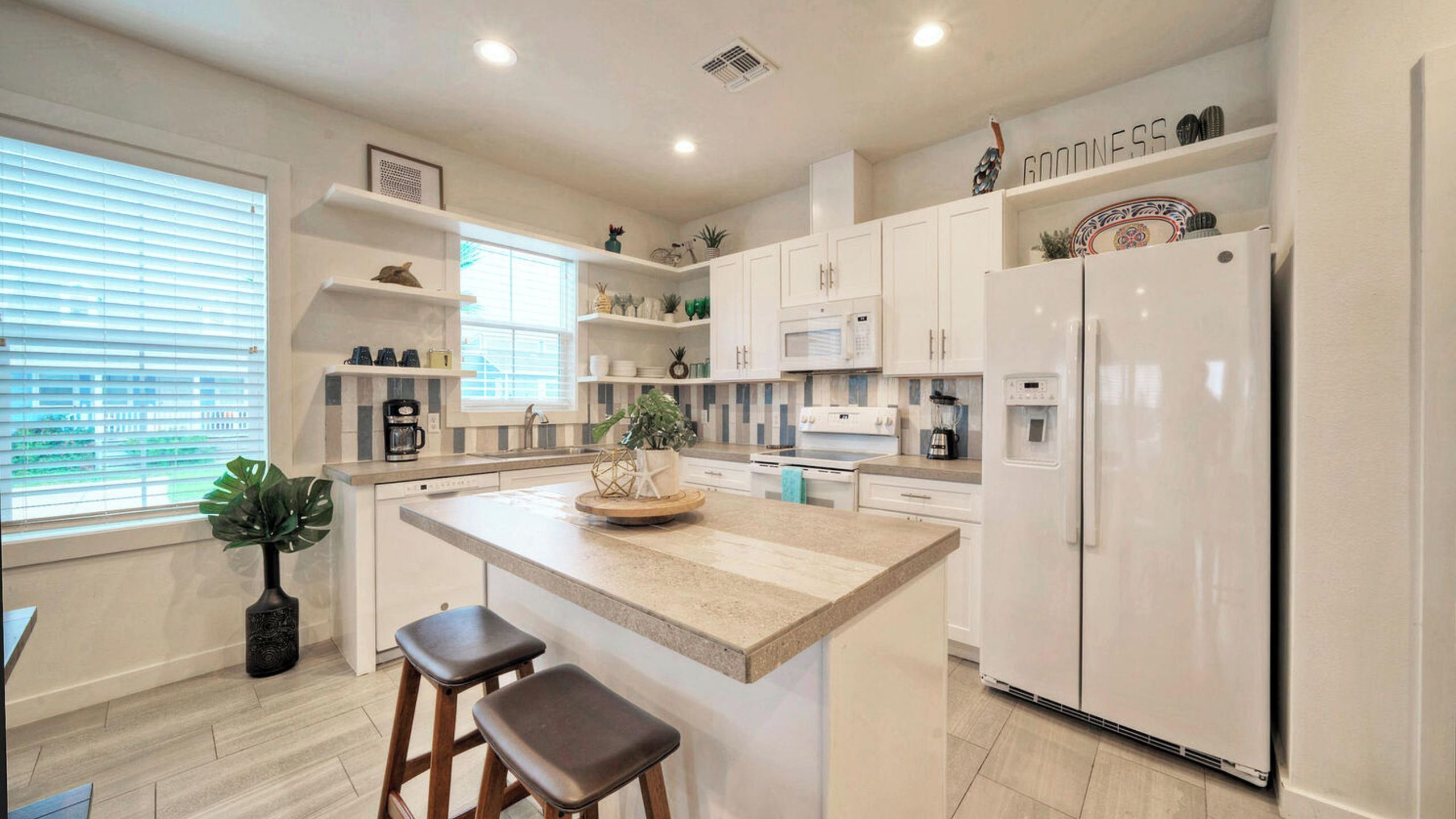 A modern kitchen with white cabinets, a central island, bar stools, a refrigerator, and a window with blinds.