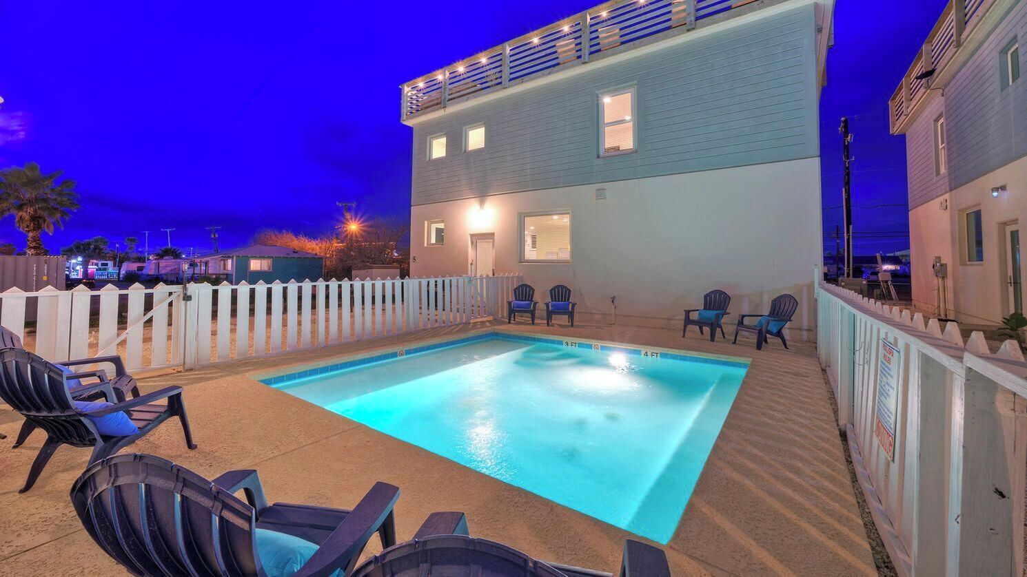 Une piscine avec des chaises longues, une clôture blanche en bois et un bâtiment à deux étages doté de fenêtres.