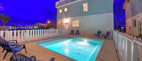 A swimming pool with lounge chairs, a white picket fence, and a two-story building with windows.