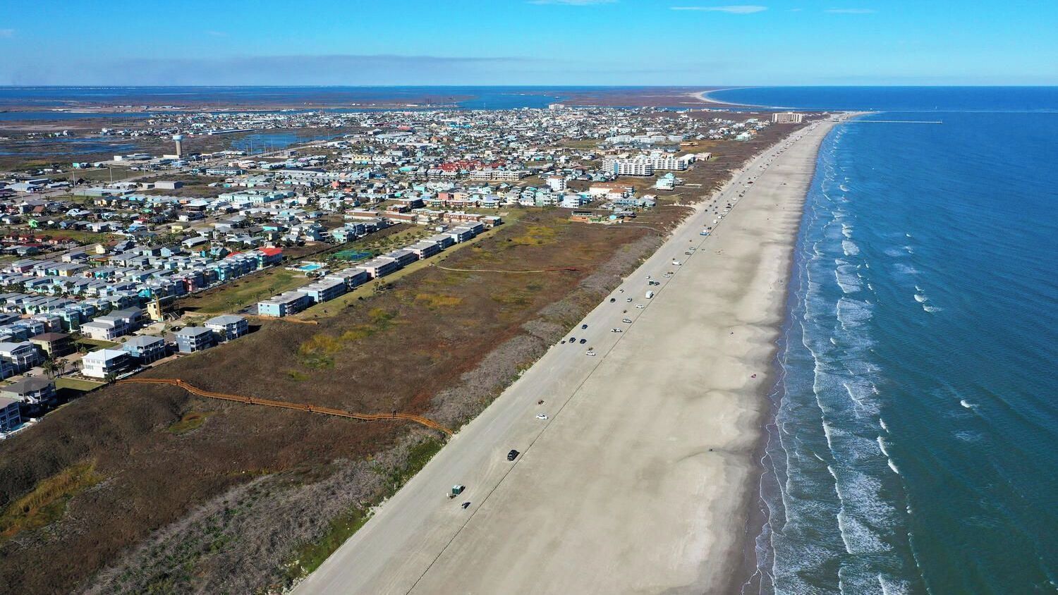 An aerial view of a coastal town with a long beach, residential buildings, and a clear blue sky.