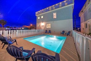 A swimming pool with lounge chairs, a white picket fence, and a building with lights.
