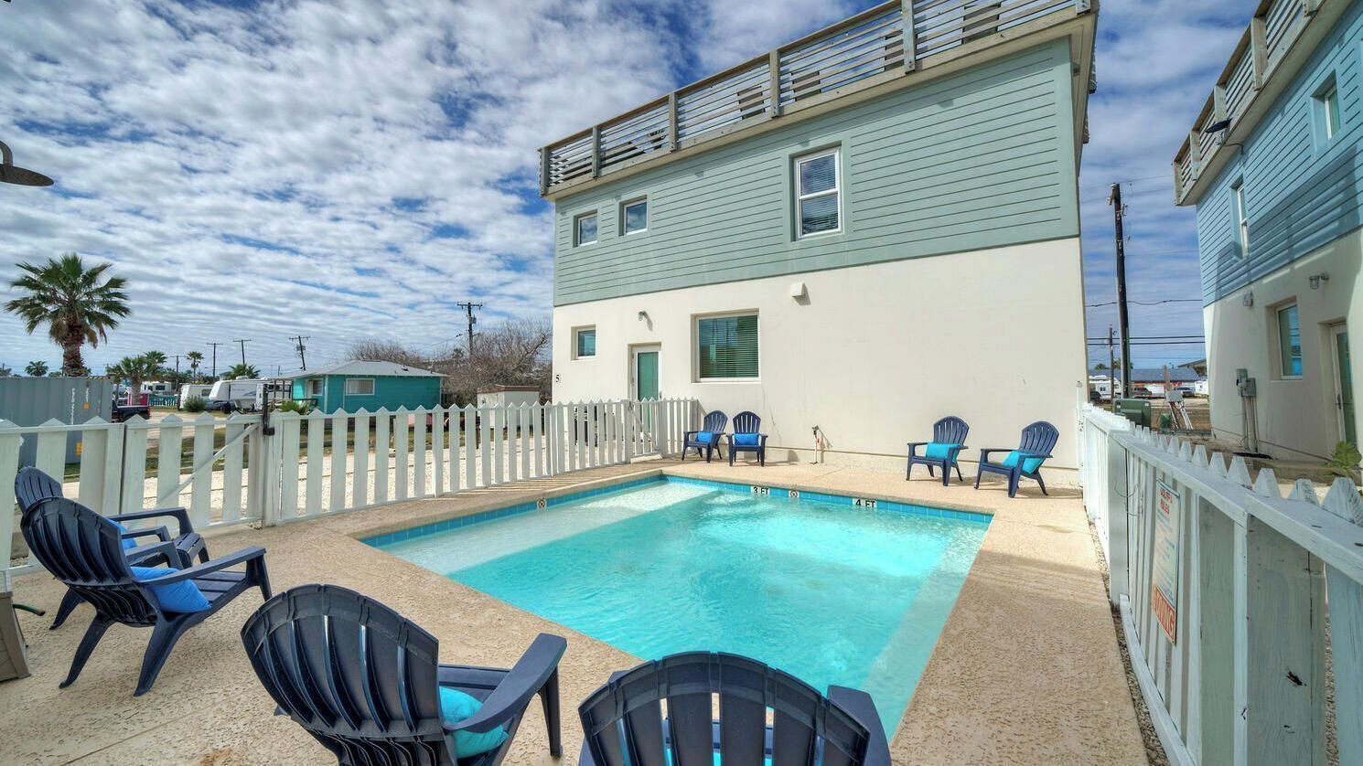 A pool area with chairs, a white picket fence, and a two-story building with a light blue exterior.