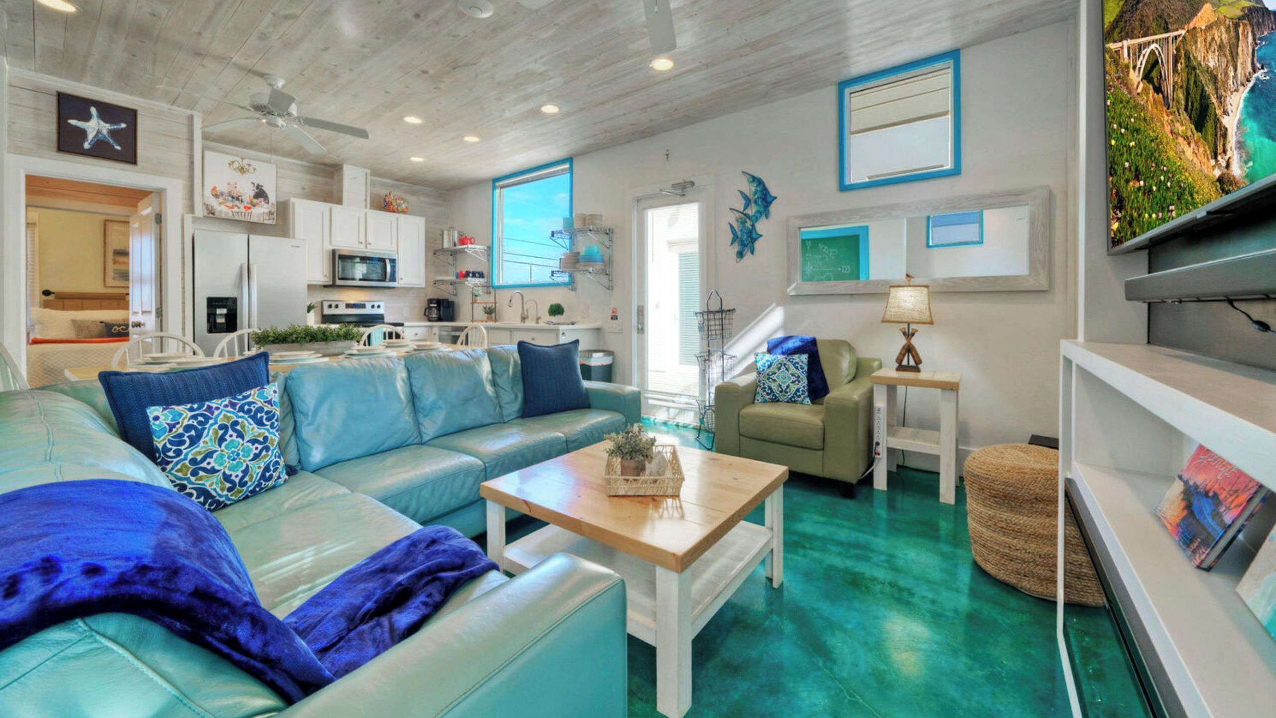 A modern living room with a teal sofa, a wooden coffee table, and a kitchen area with white cabinets and a microwave.