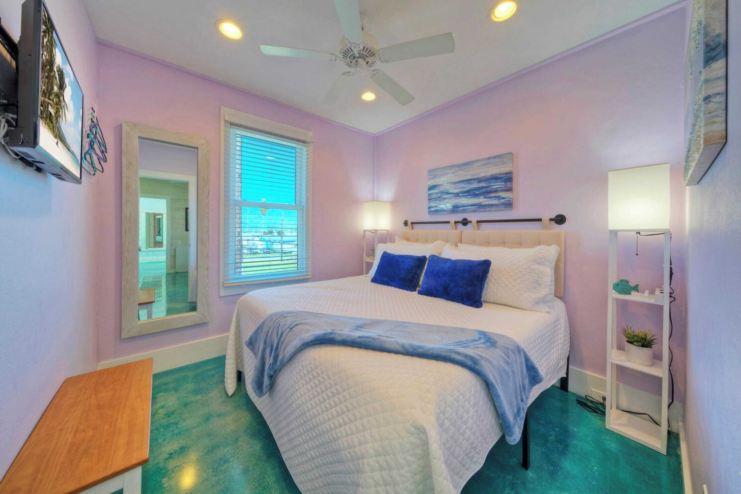 A bedroom with a bed, a ceiling fan, a window with blinds, a bedside lamp, and a small shelf with a plant.