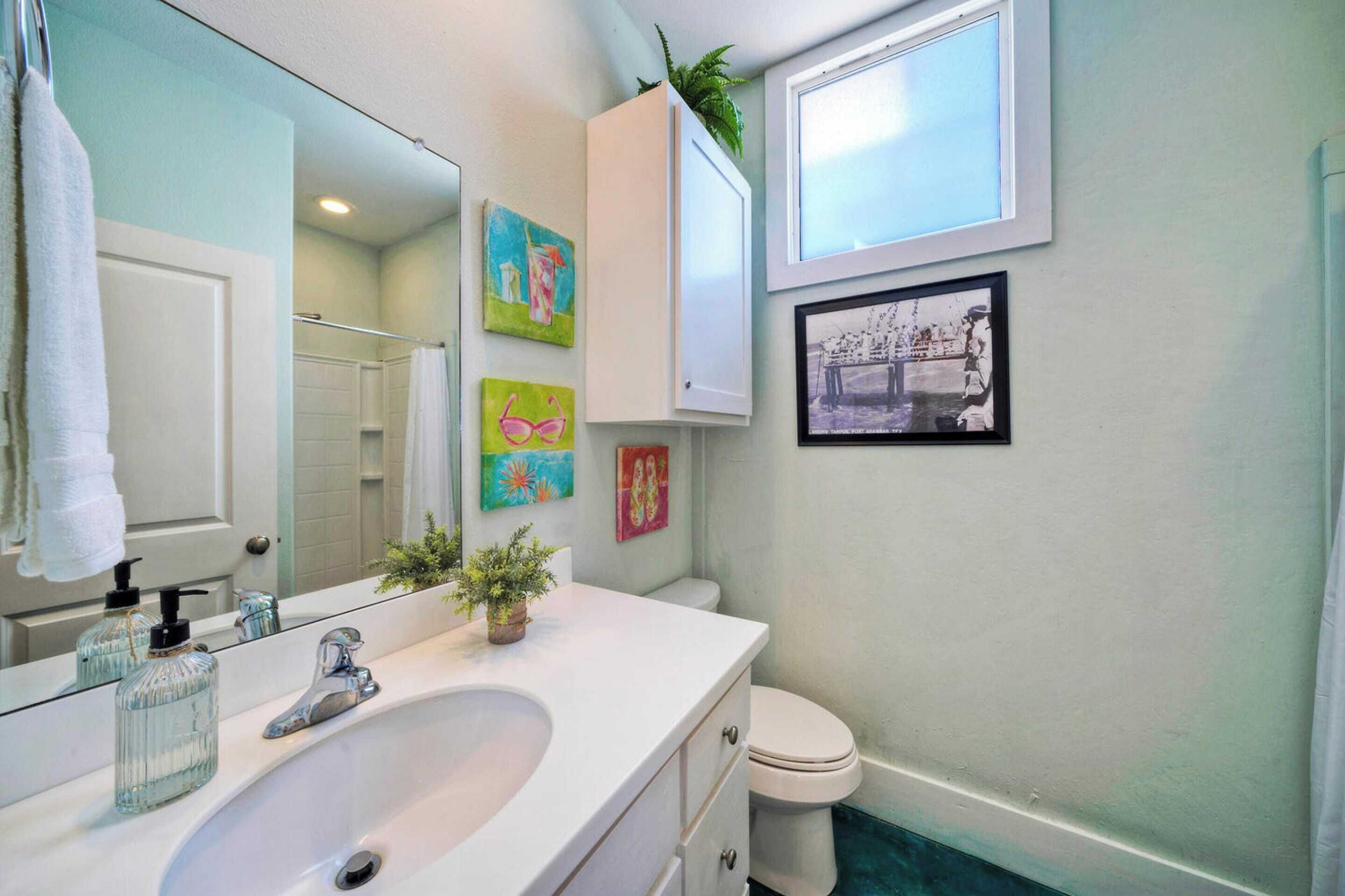 A bathroom with a white sink, a mirror, a toilet, and a window. There are three paintings on the wall and a plant on the counter.