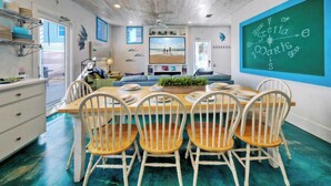 A dining area with a wooden table and white chairs, a chalkboard with writing, and a view of the beach through a window. - Stella Maris by Avantstay Rooftop View + Pool (Port Aransas)