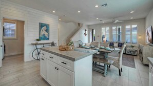 A spacious kitchen with white cabinetry, a central island, and a dining area with wicker furniture. - Breakaway by Avantstay Near Music, Eats (Port Aransas)