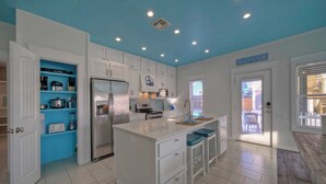 A kitchen with white cabinets, a blue accent wall, and a sign reading "BEACH HOUSE". - Pool Access, Private Patio Living, Near the Beach Seahorse Cove by Avantstay (Port Aransas)