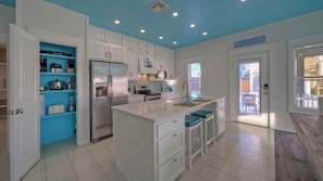 Cozinha com armários brancos, geladeira de aço inoxidável, bancada azul e placa com a inscrição 'BEACH HOUSE'.