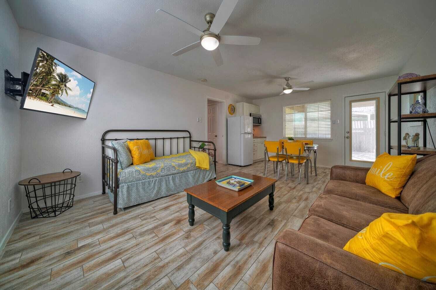 A living room with a sofa, coffee table, and a dining area with chairs. There is a kitchen area with a refrigerator and microwave.