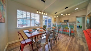 A dining area with a wooden table and metal chairs, a kitchen with white cabinets, and a red couch in the background. - Sun Beach by Avantstay Private Pool, Near Beach (Port Aransas)