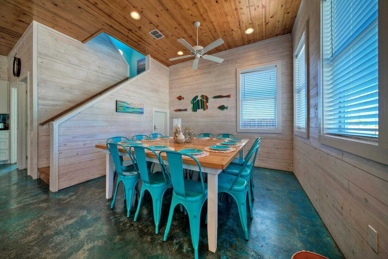 A dining area with a wooden table and turquoise chairs, a ceiling fan, and a staircase leading to an upper level.