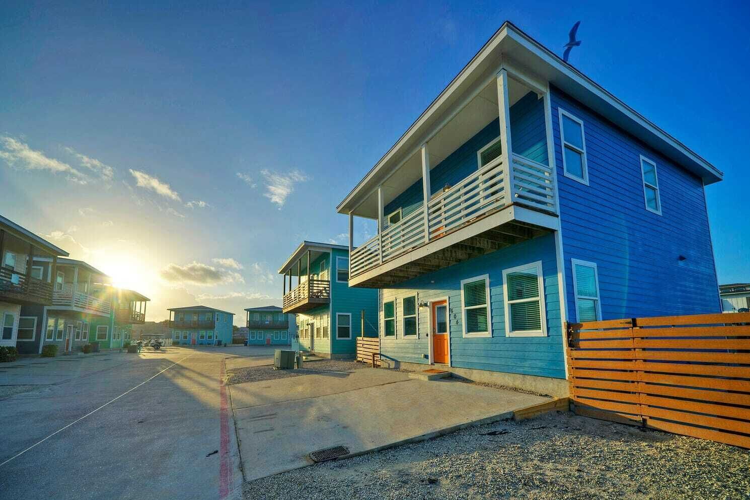 A row of blue two-story houses with balconies, a wooden fence, and a clear sky.