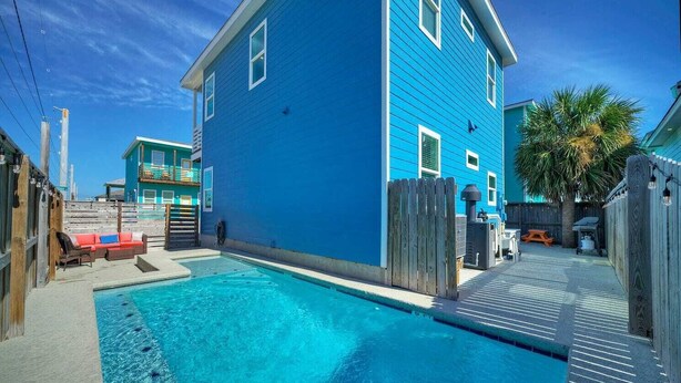 A residential area with a swimming pool, a blue house, and a wooden fence. - Private Pool+patio, Arcade Game, Walk to the Beach Ocean Devotion by Avantstay (Port Aransas)