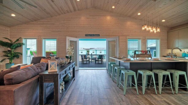 A spacious living area with a sofa, a coffee table, and a fireplace. There is a kitchen area with a bar and stools. The room has wooden floors and a high ceiling with exposed beams. - Private Pool, Covered Patio 4 Blocks to Beach Bourbon Breeze by Avantstay (Port Aransas)