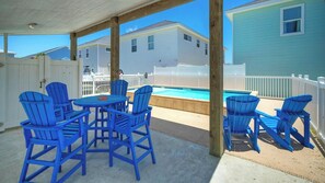 A covered patio with blue Adirondack chairs, a pool, and a white fence.