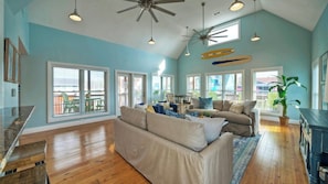 A spacious living room with a high ceiling, large windows, and a ceiling fan.