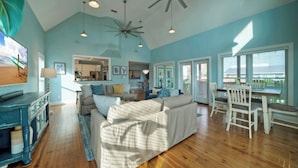 A spacious living room with a large sofa, a dining table with chairs, a ceiling fan, and a view through large windows. - Moxy's Beach House by Avantstay Pool + Views (Port Aransas)