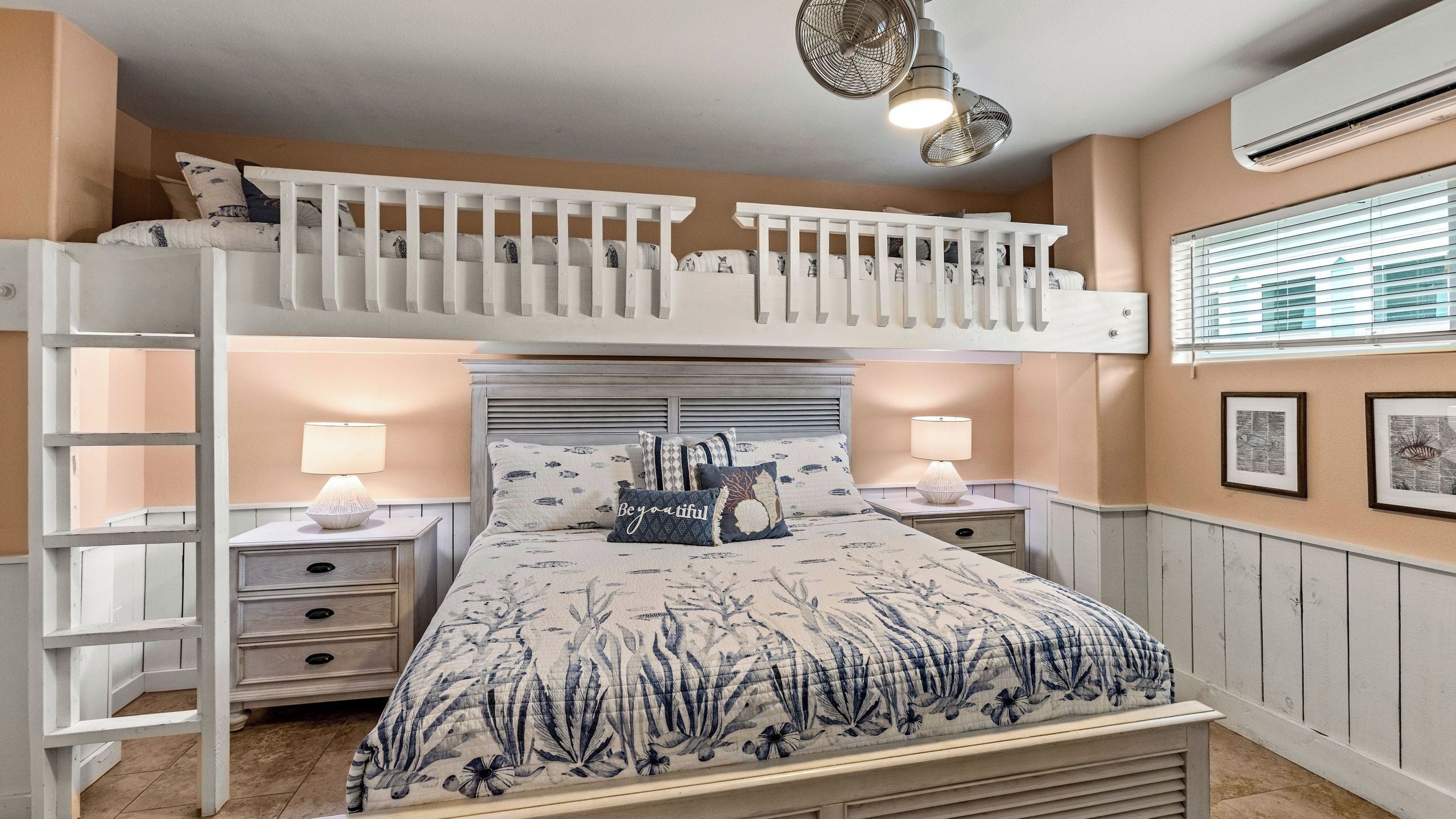 A bunk bed with a ladder, two bedside tables with lamps, a bed with floral bedding, and a window with blinds.