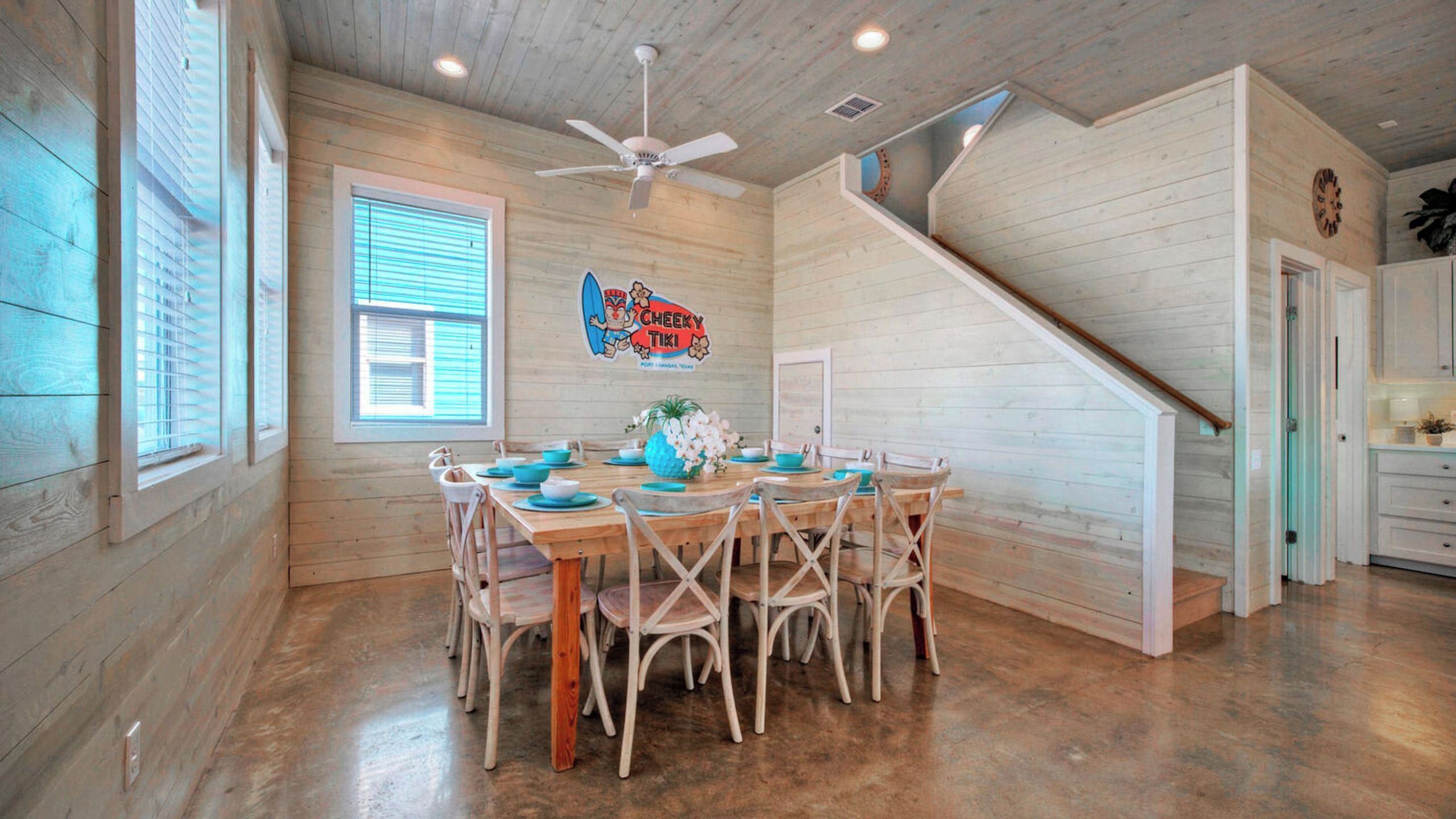 Un intérieur en bois avec une table à manger dressée pour six personnes, un ventilateur de plafond et un escalier menant à un autre niveau.