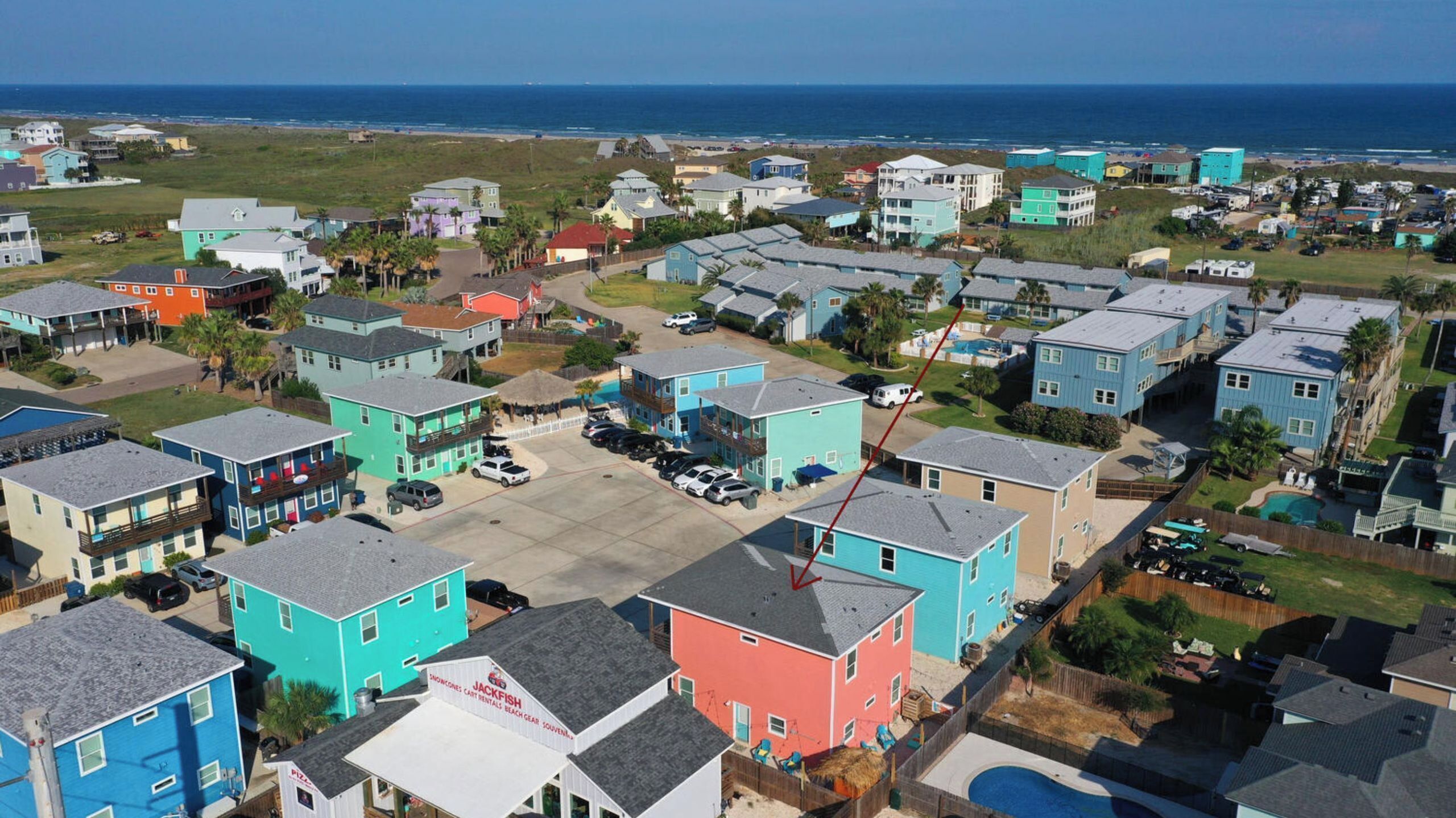 Un quartier résidentiel en bord de mer, avec des maisons aux couleurs vives, une piscine bien visible et une plage en arrière-plan.