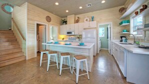 A kitchen with wooden cabinets, a central island with stools, a microwave, and a refrigerator. - Backyard Tiki Bar, Walk to the Beach, Pool Cabana Cheeky Tiki by Avantstay (Port Aransas)
