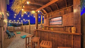 An outdoor wooden bar area with string lights, a TV screen, and wooden furniture. - Backyard Tiki Bar, Walk to the Beach, Pool Cabana Cheeky Tiki by Avantstay (Port Aransas)
