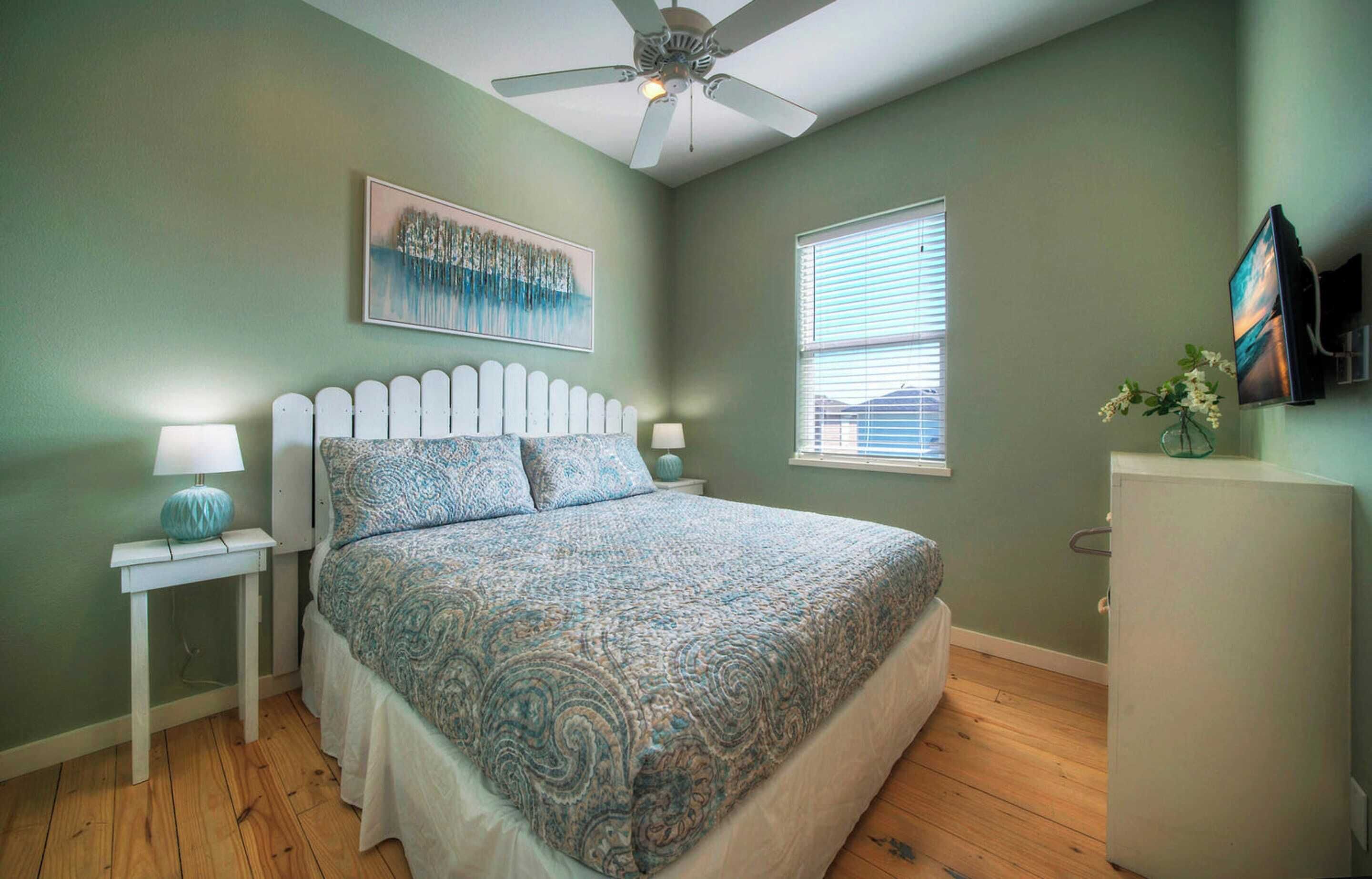 A bedroom with a bed, a ceiling fan, a window, and a TV on the wall.