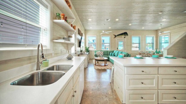 A spacious kitchen with white cabinetry, a large island, and a dining area with a green sofa. - Backyard Tiki Bar, Walk to the Beach, Pool Cabana Cheeky Tiki by Avantstay (Port Aransas)
