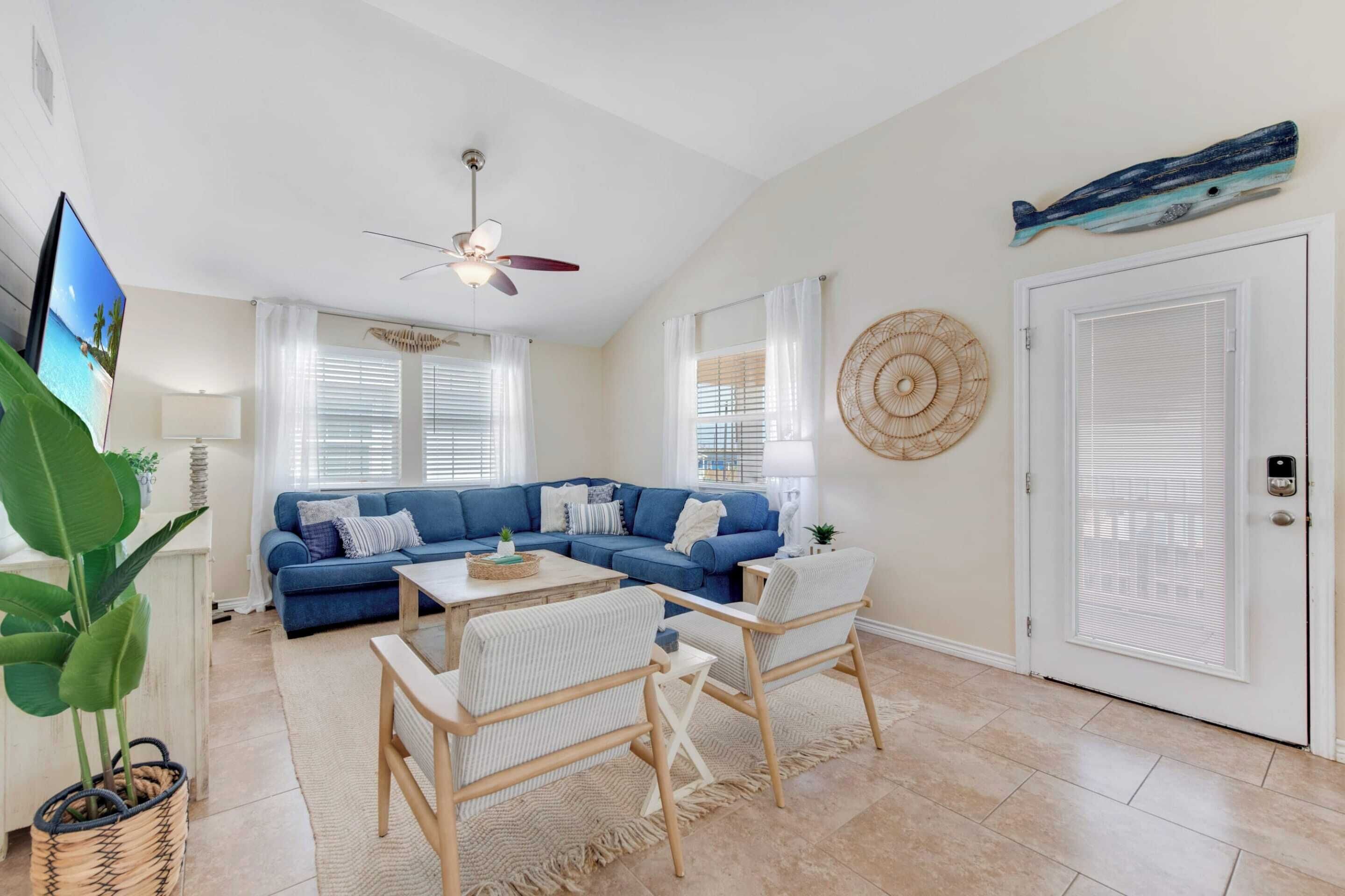 A living room with a blue sofa, a coffee table, and two chairs. There is a ceiling fan, a TV, and a door with a window.