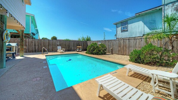 Une piscine rectangulaire dotée d’une terrasse en bois et de chaises longues, entourée d’une clôture en bois et de verdure.