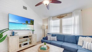 Een woonkamer met een televisietoestel dat een strandscène toont, een blauwe bank, een houten salontafel en een plafondventilator.