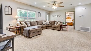 A spacious living room with a large L-shaped sectional sofa, a wooden coffee table, and a ceiling fan. There is a door leading to a bedroom with a bed and a nightstand. - Rural Retreat w/ Covered Porch Near Jackson (Grove Hill)
