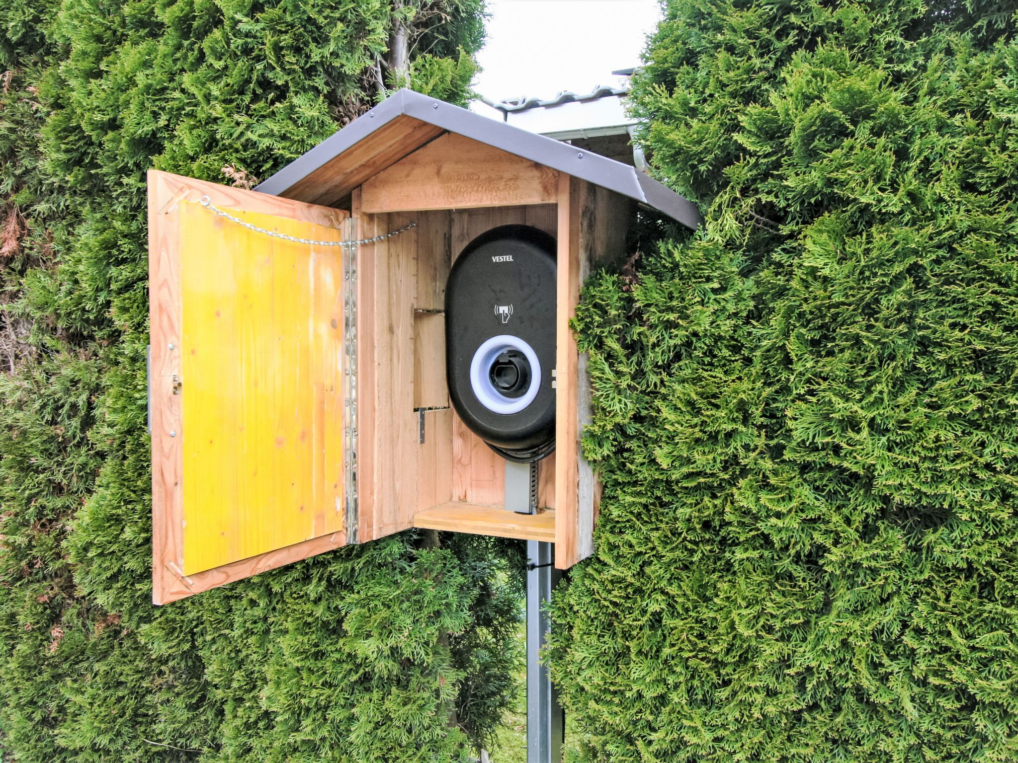 Un comedero de madera con puerta amarilla y una cámara negra en su interior, rodeado de follaje verde y frondoso.