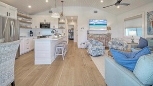 A spacious kitchen with white cabinets, a central island, and bar stools. There is a living area with a sofa and a television mounted on the wall. - Boujee Pelican by Avantstay Pool+game Room+views (Port Aransas)