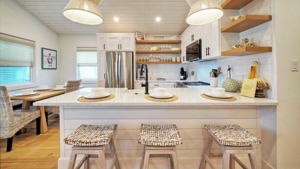 A modern kitchen with white cabinetry, a central island with stools, and pendant lighting. - Boujee Pelican by Avantstay Pool+game Room+views (Port Aransas)