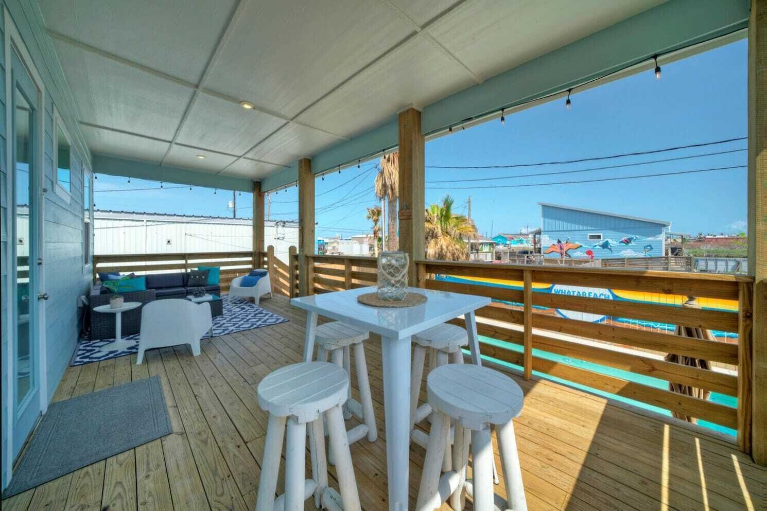 Un espace extérieur abrité doté d’une terrasse en bois, de mobilier blanc et offrant une vue sur une plage et un parc d’attractions en arrière-plan.