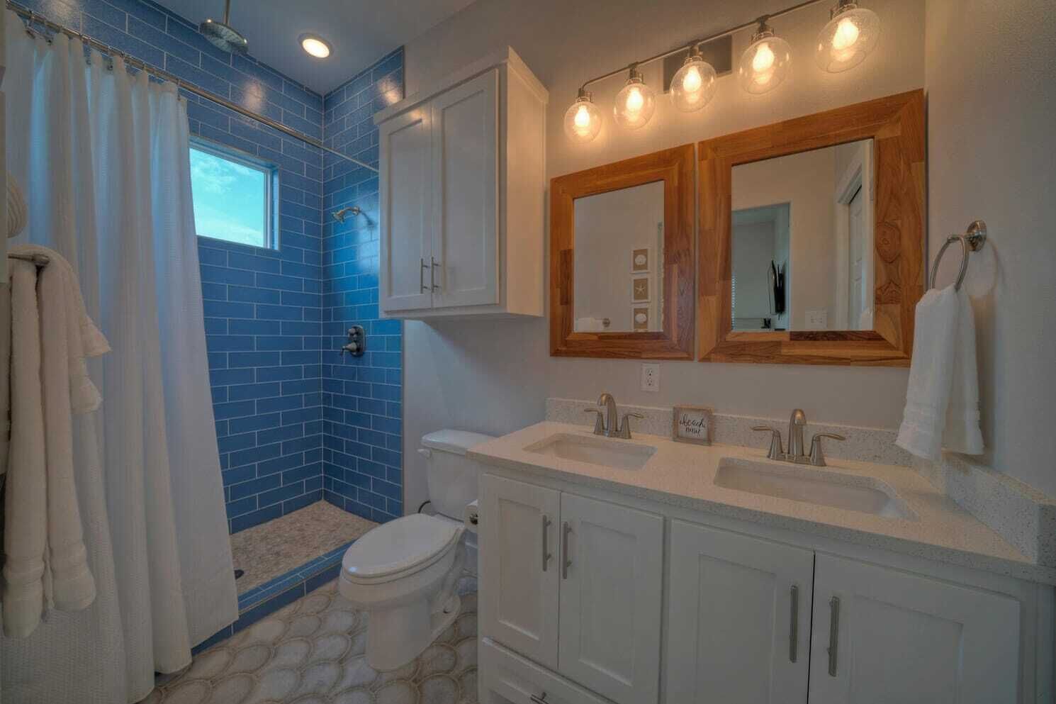 Un baño con paredes de azulejos azules, inodoro blanco, zona de ducha y tocador doble con espejo.