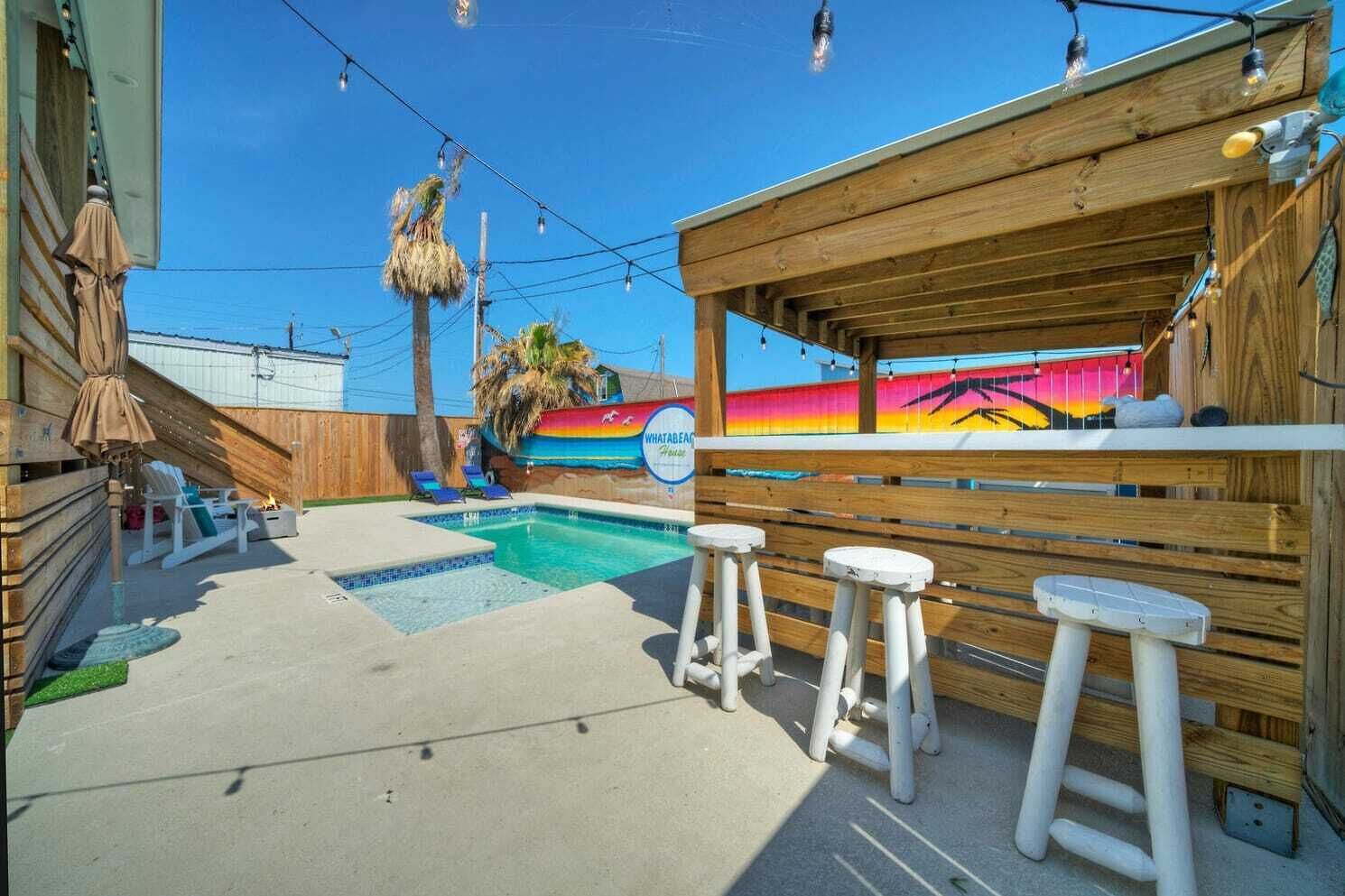 Een achtertuin met een zwembad, een houten terras, lounge stoelen en een bar.