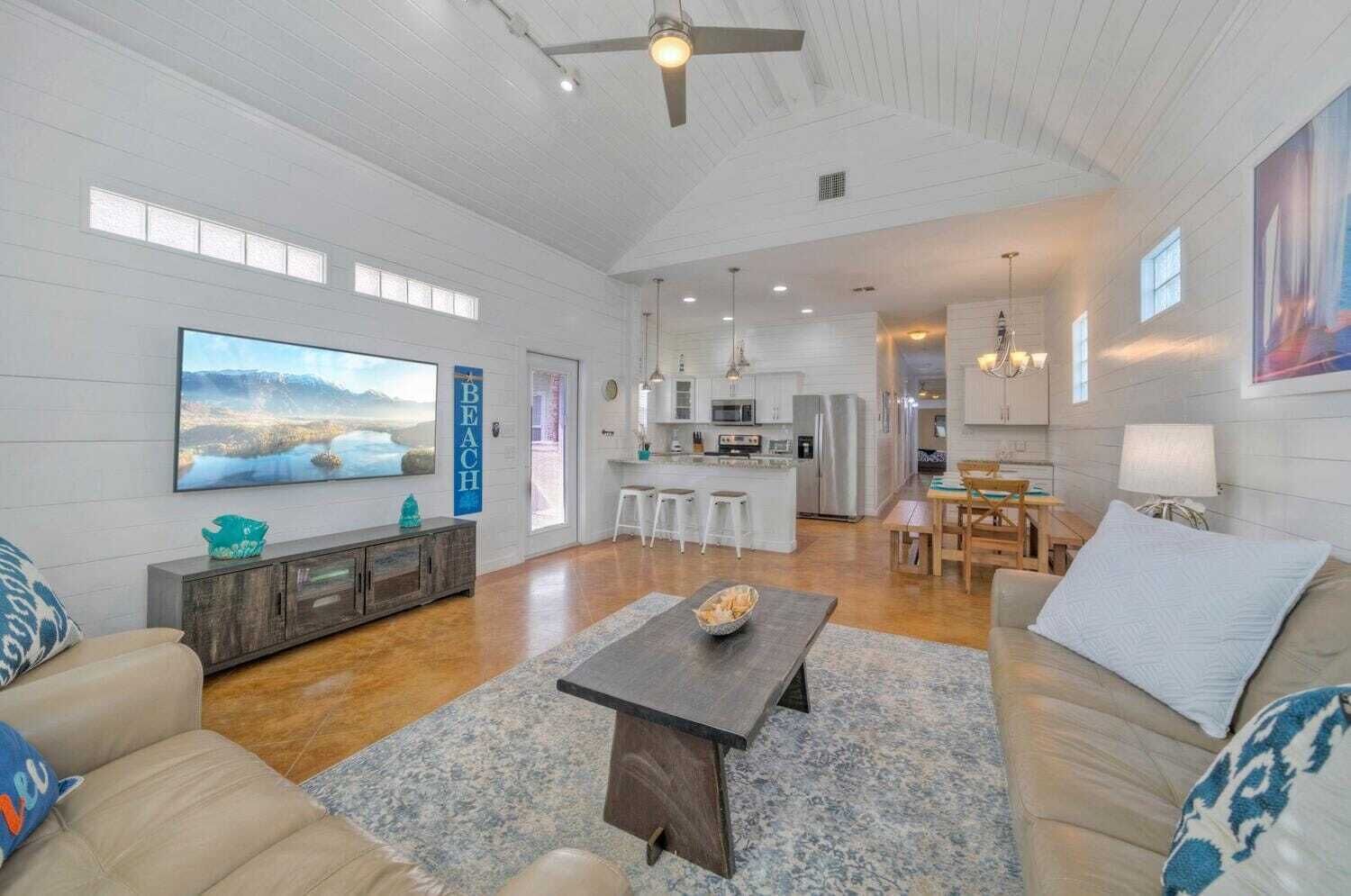A spacious living room with a large TV, a dining area, and a kitchen in the background.