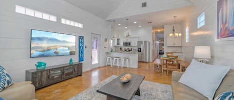 A spacious living room with a large TV, a dining area, and a kitchen in the background.
