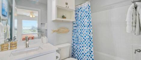 Un baño con lavamanos, inodoro y ducha blancos. Hay una cortina de ducha azul y blanca, un mueble blanco sobre el lavamanos y una planta pequeña en el mueble.