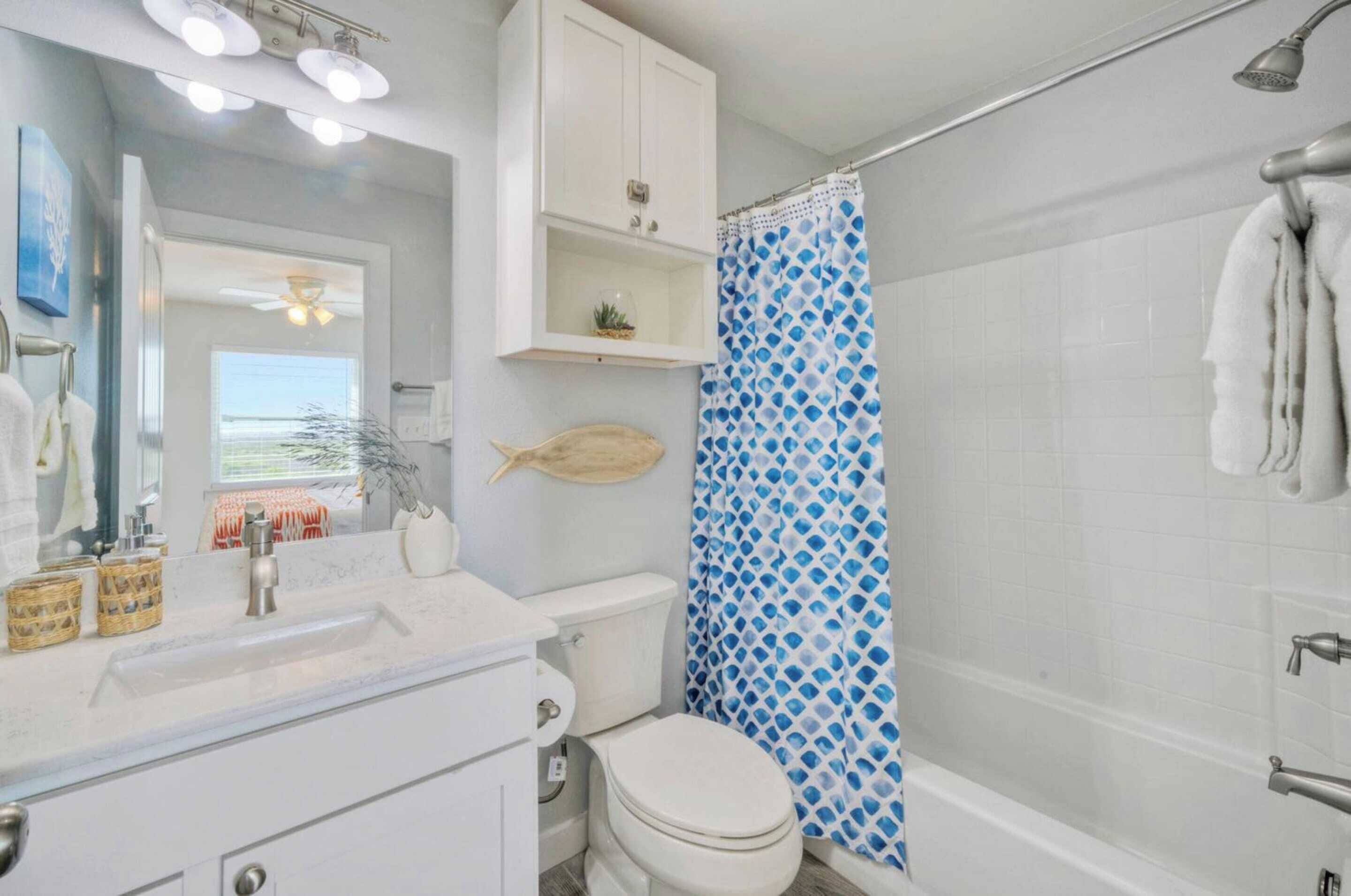 Un baño con lavamanos, inodoro y ducha blancos. Hay una cortina de ducha azul y blanca, un mueble blanco sobre el lavamanos y una planta pequeña en el mueble.